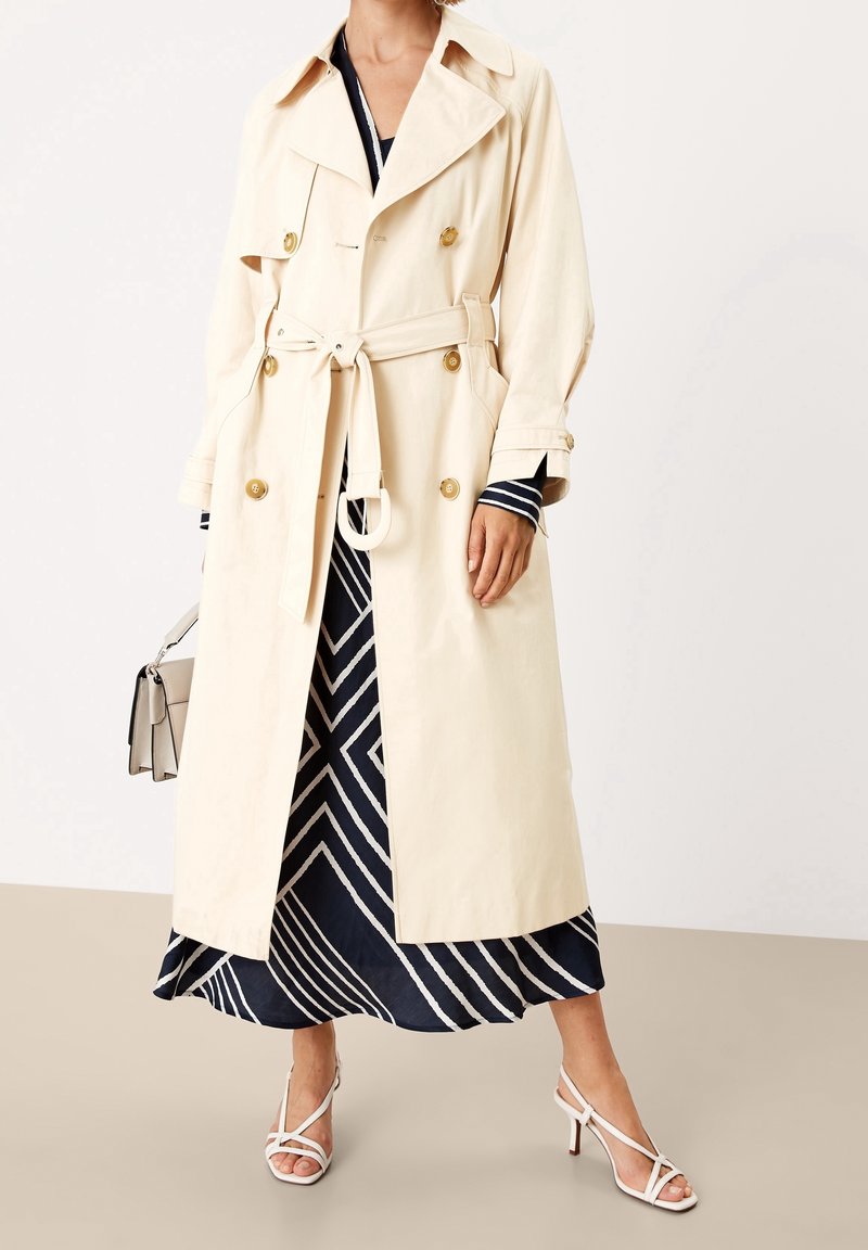 Gabardina beige con botones cruzados, cinturón en la cintura y ajuste holgado. Lleva puesta sobre un vestido azul marino con estampado y zapatos de tacón blancos.