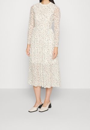 Kvinde iført en hvid midikjole med lange gennemsigtige ærmer og flerfarvet lille blomsterprint, kombineret med cremefarvede lave blokhælede slingback-sko.