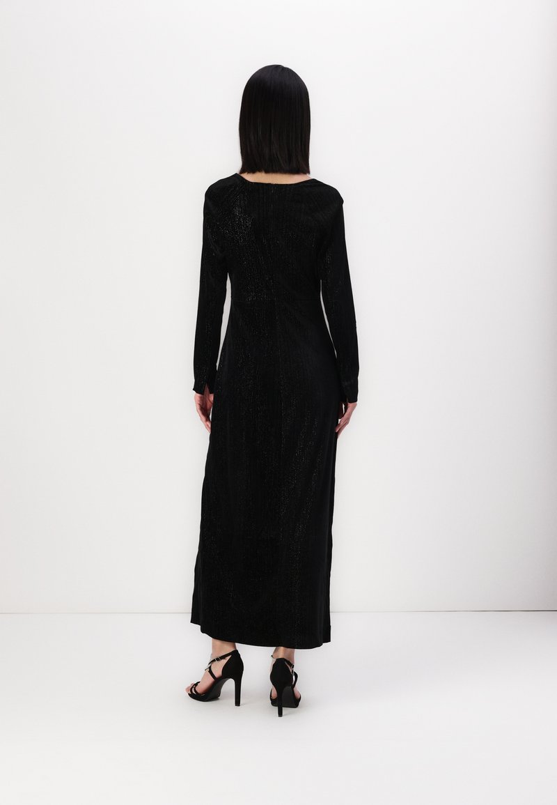 Langes schwarzes Kleid mit einer eleganten, taillierten Silhouette, langen Ärmeln und texturiertem Stoff. Kombiniert mit schwarzen High Heels.