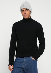 Fekete garbó puha anyagból, testhez simuló szabás, bordázott mandzsettákkal. Kék farmernadrággal és világosszürke kötött sapkával viselve.