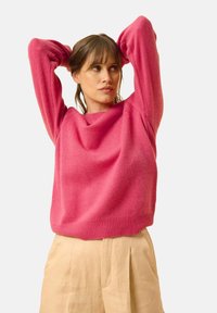 Pullover rosa lavorato a maglia con una vestibilità morbida, scollatura rotonda e polsini a coste. Il modello indossa pantaloni beige a pieghe.