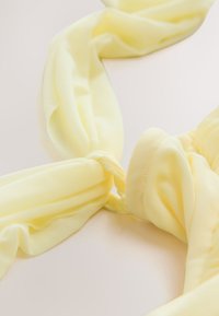 Pehme vaaleankeltainen kangas, joka on kieritetty ja solmittu vaalealle pinnalle ja näyttää tekstuuria ja lempeitä laskoksia.