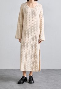 Persson bär en krämfärgad lång kofta med flätad struktur och sidslits, samt svarta snörskor på ett grått golv.