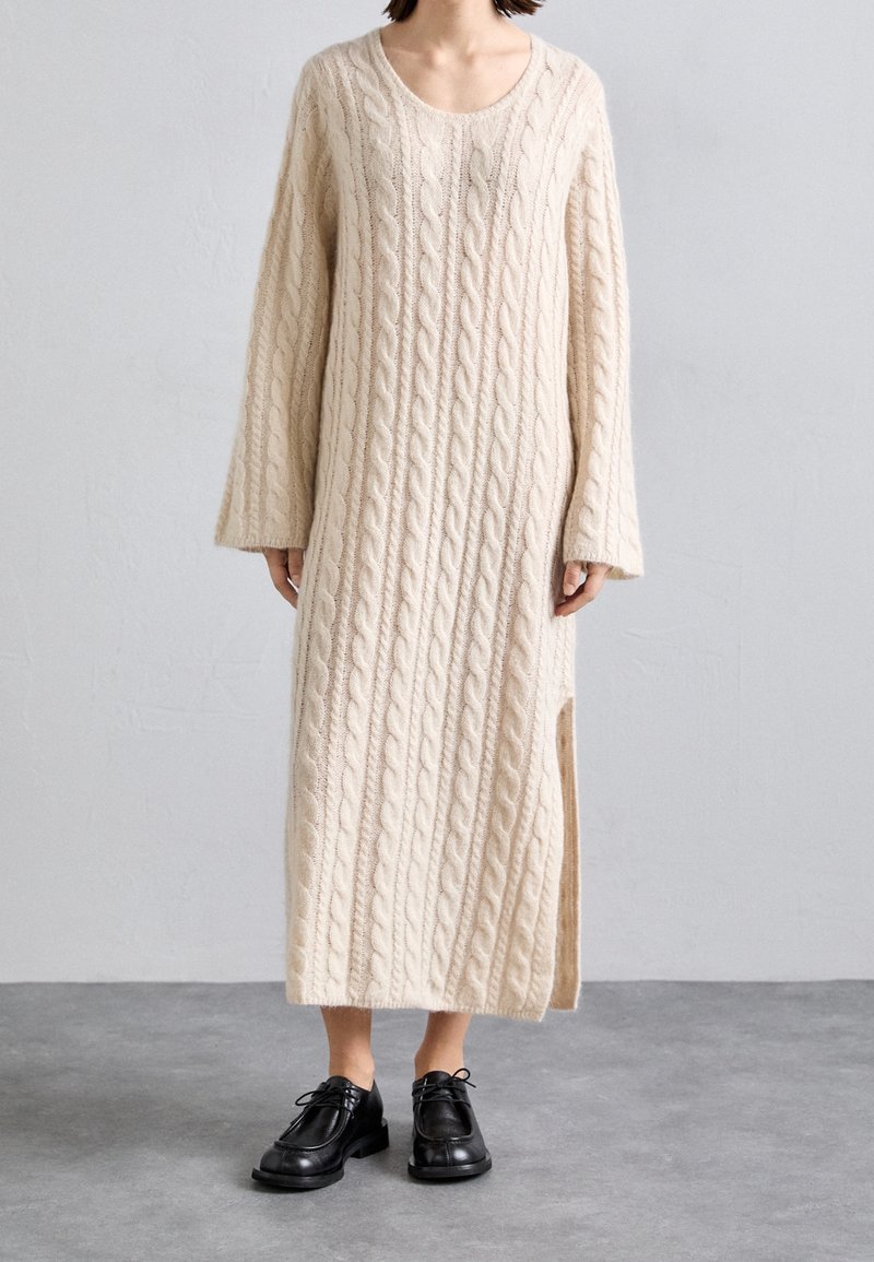 Persson bär en krämfärgad lång kofta med flätad struktur och sidslits, samt svarta snörskor på ett grått golv.