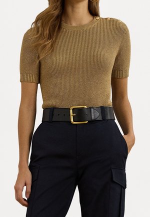 Femme portant un top marron côtelé à manches courtes avec des boutons dorés à l'épaule, une ceinture noire avec une boucle dorée, et un pantalon foncé avec des poches.
