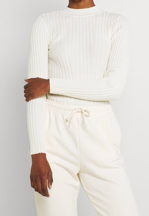Osoba w białym prążkowanym swetrze z długim rękawem i białych spodniach z wiązaniem, z jedną ręką skrzyżowaną na ciele.