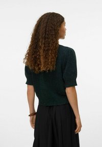 Pull en maille vert foncé à manches courtes et coupe raccourcie, avec un tissu texturé. Porté avec une jupe noire fluide.