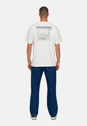 Weißes Baumwoll-T-Shirt mit einer Linienzeichnung eines Cafés auf der Rückseite. Kombiniert mit dunkelblauen Jeans und Turnschuhen.