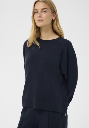 Jeune femme aux longs cheveux blonds portant un sweat-shirt bleu marine uni foncé avec des fermetures éclair latérales, debout sur un fond clair.