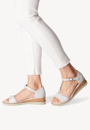 Szare sandały z odkrytymi palcami, z paskiem w kształcie litery T, koturnem owiniętym korkiem oraz teksturowanymi zdobieniami na wierzchu. Noszone z białymi spodniami.