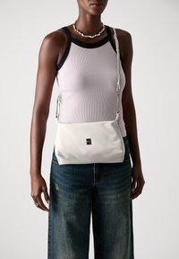Hvit skinn crossover bag med teksturert overflate, rektangulær form, justerbar stropp og en liten svart logo foran.