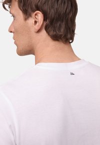 Biała koszulka z okrągłym dekoltem wykonana z bawełny. Ma małe czarne logo w pobliżu dekoltu z tyłu. Gładka tekstura i prosty design.