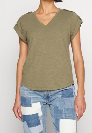 Kvinde iført olivengrøn V-hals kortærmet skjorte med opkrullede ærmer og lapper på blå jeans mod ensfarvet baggrund.