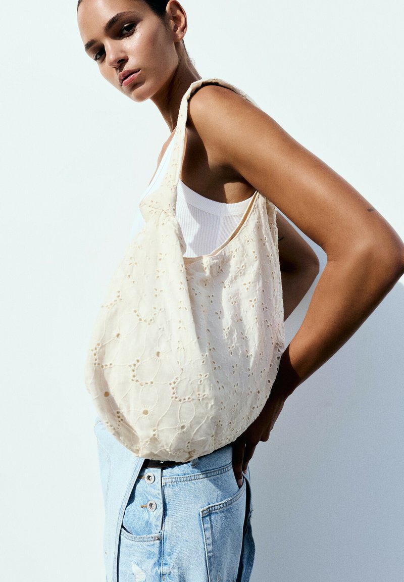 Femme portant un débardeur blanc et un jean bleu clair porte un grand sac en tissu à œillets floraux couleur crème sur l'épaule.