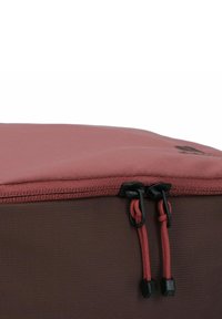 Bolsa de tela roja con una superficie texturizada, con una cremallera parcialmente cerrada y dos cuerdas negras. Panel inferior marrón visible.