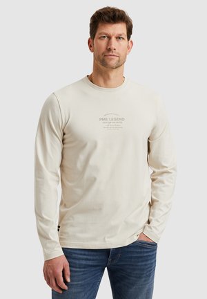 Long-sleeve beige cotton t-shirt featuring a crew neck and front graphic text. Paired with blue jeans, shown on a neutral background.