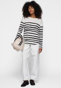 Gestreiftes langärmliges Oberteil in Off-White und Schwarz, kombiniert mit hellgrauen Jogginghosen und hält eine beigefarbene Daunenjacke. Schwarze Schuhe runden das Outfit ab.