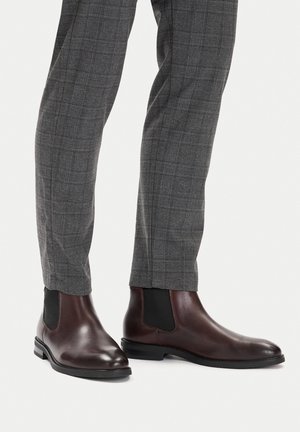 Rudos odinės Chelsea batai su elegantišku dizainu, elastingomis šoninėmis skydeliais ir apvalia nosimi, derinami su pilkais languotais siaurėjančiais kelnėmis.