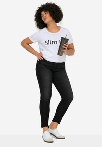 Balta trumpa marškinėliai su užrašu „Slim Fit“, juodos džinsinės kelnės su išplyšusiu kraštu ir baltos sportbačiai su pilkais akcentais. Laiko juodą šiaudinį puodelį.