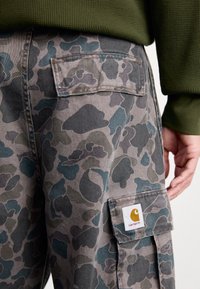 Spodnie cargo w kamuflażu w stonowanej szarości z ciemnozielonymi akcentami, z kieszeniami łatanymi i logo Carhartt na prawej nodze.