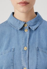 Ljusblå denimskjorta med krage, tre knappar och en bröstficka. Slät textur med klassisk passform och enkel design.
