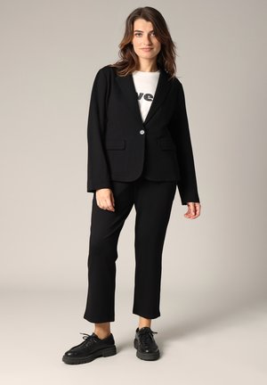 Zwarte blazer met geknipte revers en één knoop, gecombineerd met zwarte broek en zwarte veterschoenen. Witte graphic T-shirt eronder.