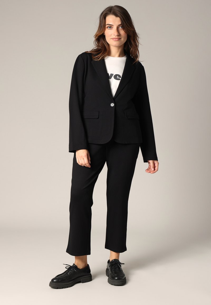 Blazer noir à un bouton, associé à un pantalon noir et un t-shirt graphique blanc, complété par des chaussures à lacets noires.