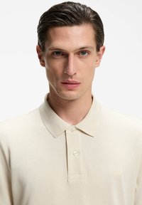 Hellbeiges Polohemd aus strukturiertem Stoff mit Kragen und Zweiknopf-Leiste. Verfügt über ein dezentes "BOSS"-Logo auf der Brust.