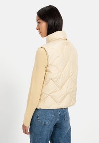 Lys gul, quiltet vest med høj krave og diagonale syninger, lavet af skinnende materiale. Båret over en beige langærmet bluse.