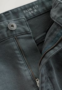 Pantalones ajustados de tela gris oscuro con una cremallera y botón visibles. La etiqueta muestra la talla 34 R. Presentan una textura suave y costuras detalladas.