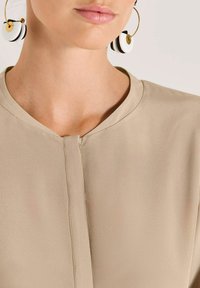 Beige Bluse mit rundem Ausschnitt und einer Knopfleiste vorne. Besitzt eine zarte Stofftextur. Schwarz-weiße, kreisförmige Ohrringe schaffen einen Kontrast.
