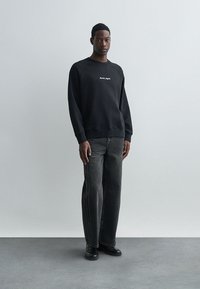 Hanorac negru din bumbac cu text alb, asortat cu blugi gri închis ușor largi, cu cusături laterale, purtat cu pantofi negri.