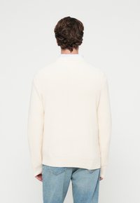 Κρεμ ψηλόμεσο πλεκτό πουλόβε�ρ με ραβδωτή υφή, άνετη εφαρμογή και V-σχήματος λαιμόκοψη, φορεμένο πάνω από ένα ανοιχτό μπλε πουκάμισο με γιακά.