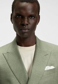 Lichtgroene blazer met textuur en een getailleerde pasvorm, gecombineerd met een crèmekleurig overhemd. Witte pochet in de voorzak van de blazer. Close-up view.