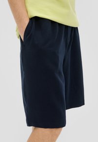 Marineblaue Shorts aus weichem Stoff mit einem elastischen Bund und seitlichen Taschen, teilweise von einem hellgrünen Oberteil bedeckt.