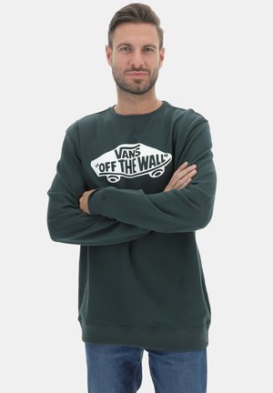 Felpa verde con una grafica del logo bianco "OFF THE WALL" di Vans. Polsini e orlo a coste, design con scollatura rotonda, abbinata a jeans blu.