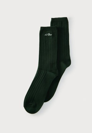 Par de calcetines acanalados de color verde oscuro con el logo "Les Deux" bordado en blanco cerca del puño superior, mostrados planos sobre un fondo claro.