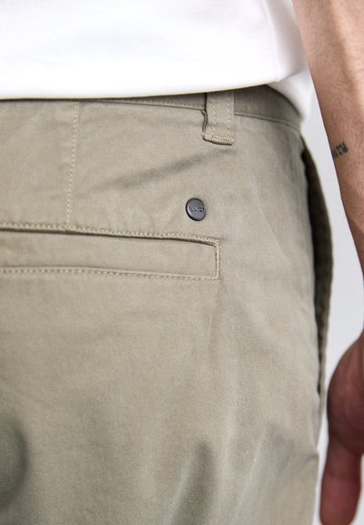 Pantalons en coton olive, dotés d'une poche arrière avec un bouton noir, de coutures visibles et d'une texture lisse et mate.