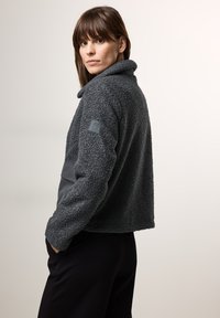 Veste en polaire gris foncé avec un col haut et une surface texturée, présentant un panneau inférieur noir et un patch logo sur la manche.