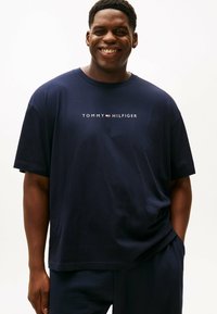 Uśmiechający się mężczyzna w granatowej koszulce Tommy Hilfiger z logo tekstowym na środku klatki piersiowej, stojący z jedną ręką w kieszeni na białym tle.