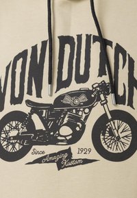 Haut à capuche beige avec un graphisme de moto noir, comprenant un œil ailé, et du texte « Von Dutch » en gras en haut, suivi de « Since 1929 Amazing Kustom » en bas.