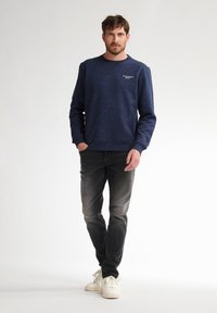 Dunkelblauer Pullover mit gesprenkelter Textur, Rundhalsausschnitt und Logodetail; kombiniert mit ausgebleichten schwarzen Jeans und weißen Sneakers.