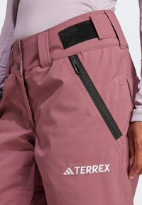 Terrex pink ski pants feature a high waistband, zippered pocket, and black Velcro accent. Made from durable material with a smooth texture.