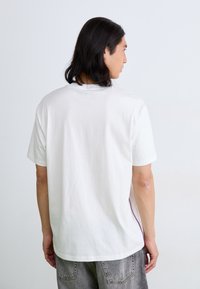 Hvid bomulds-T-shirt med korte ærmer, set bagfra. Har en afslappet pasform og en accentfarve langs sidesømmene. Enkel tekstur.