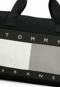 Sort stof trænings- eller rejsetaske med et hvidt og gråt panel. Fremtrædende "TOMMY JEANS" tekst i hvid på et sort bånd. Dobbelt håndtag til bæring.
