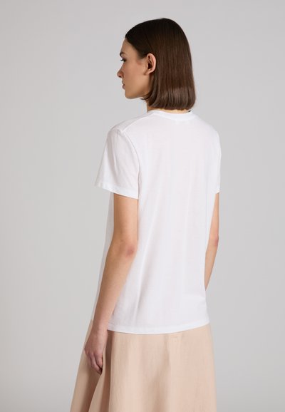 Mujer de cabello corto y castaño mirando de perfil, vestida con una camiseta blanca de manga corta lisa y una falda beige, sobre un fondo gris.