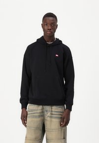 Čierna mikina s kapucňou a predným vreckom, so malou červenou nášivkou. Nosí sa s svetlými štruktúrovanými cargo nohavicami.