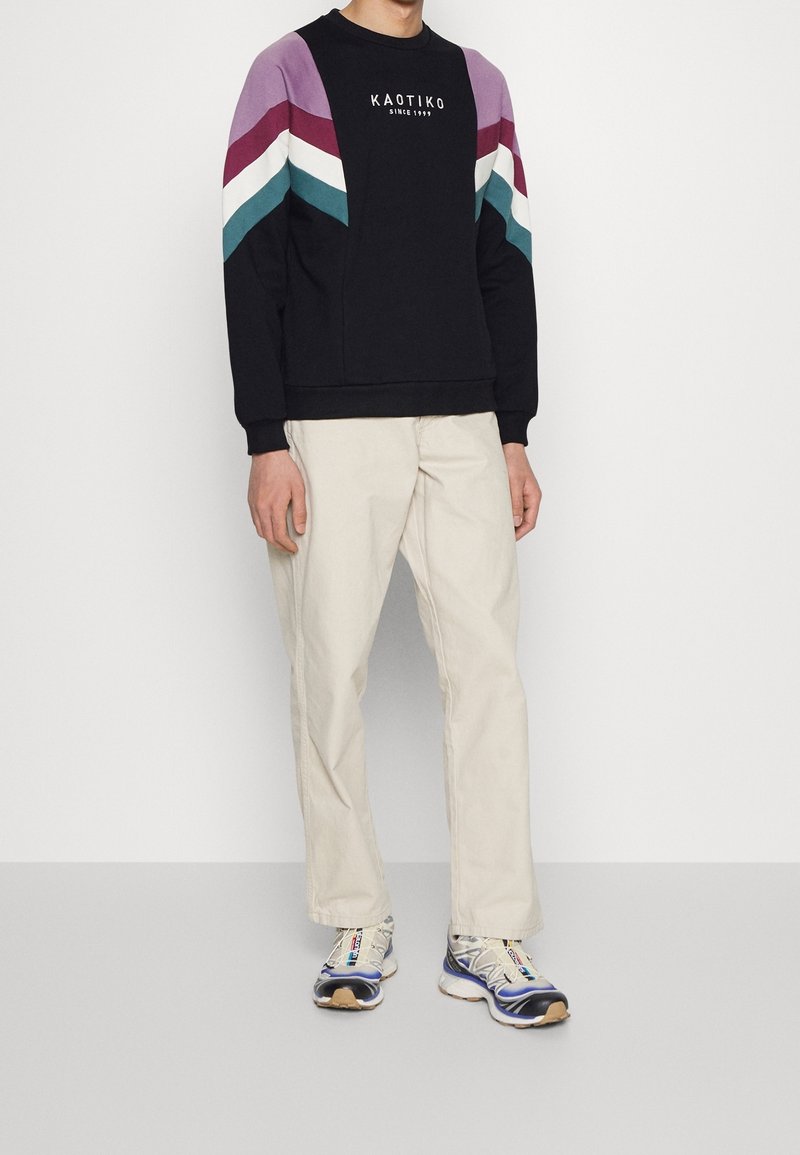 Personne portant un sweat-shirt noir avec des rayures en chevrons colorées et du texte, un pantalon beige et des baskets blanches, debout sur un fond gris.