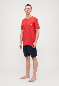 Červené bavlnené tričko s krátkymi rukávmi a bielym logom, skombinované s námorníckymi šortkami. Model stojí naboso na jednofarebnom pozadí.