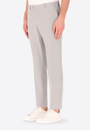 Uomo con pantaloni grigio chiaro slim fit e sneakers bianche, in piedi davanti a uno sfondo semplice con le mani rilassate lungo i fianchi.